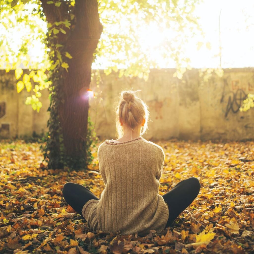 A woman sits peacefully on a rocky cliffside at sunset, meditating with her eyes closed, surrounded by nature. Her phone rests beside her, symbolizing a digital detox and Meditation to inner peace.