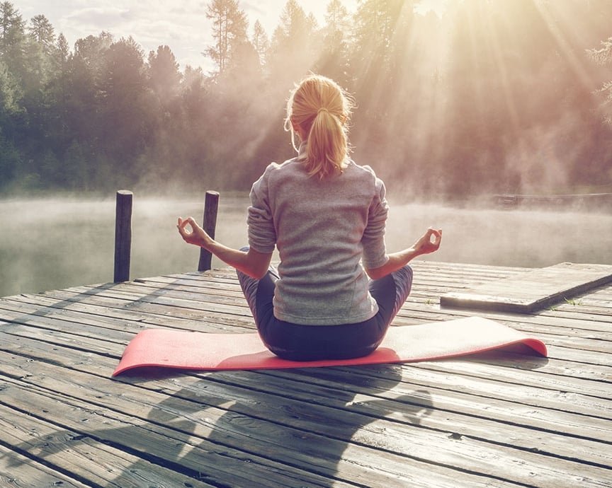 A person meditates by a calm lake surrounded by mountains and trees, embracing solitude and mindfulness during a digital detox retreat.