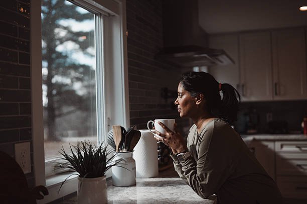 A woman taking a mindful moment as part of her mental wellness routine, holding a warm drink and looking out her kitchen window.