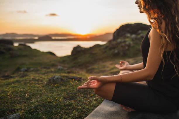 Blog Side view of a woman with long hair sitting cross-legged on a yoga mat outdoors. Her hands are in a mudra pose as she meditates at sunset with a calm lake and hills in the background.