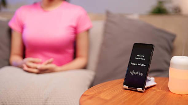 An Asian woman meditates peacefully on a yoga mat in a sunlit room, wearing headphones and using a 'Forest Whisper' natural sound app on her smartphone placed in front of her.