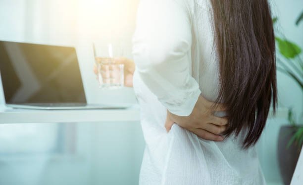 Blog Woman experiencing chronic back pain at work, holding her lower back while standing by her desk in a sunlit office."