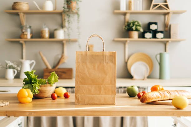Blog Illustrating a core part of sustainable living, a reusable paper grocery bag overflows with colorful, fresh vegetables and a baguette on a bright kitchen table.