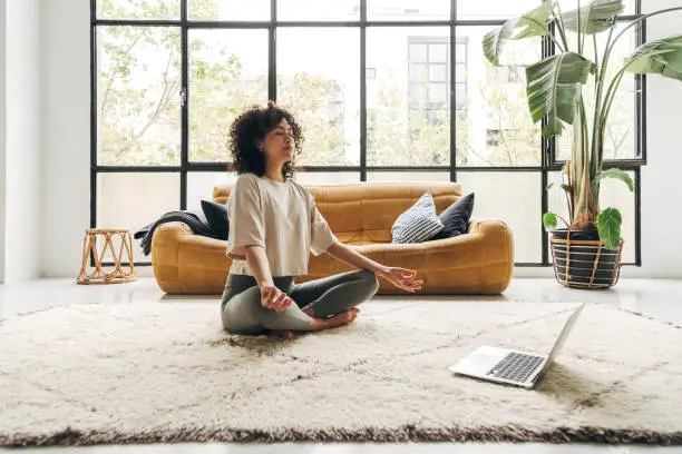  young Latina woman practicing a guided meditation for mental wellness at home, using an online video lesson on her laptop in a bright living room.