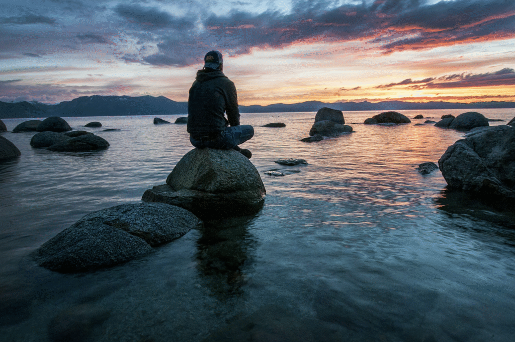 Sustainable Living: 9 Powerful Ways It Boosts Your Mental Wellness “Silhouetted figure sitting quietly on a rock at the water’s edge during sunset, symbolizing sustainable living, peace, reflection, and connection with nature.”