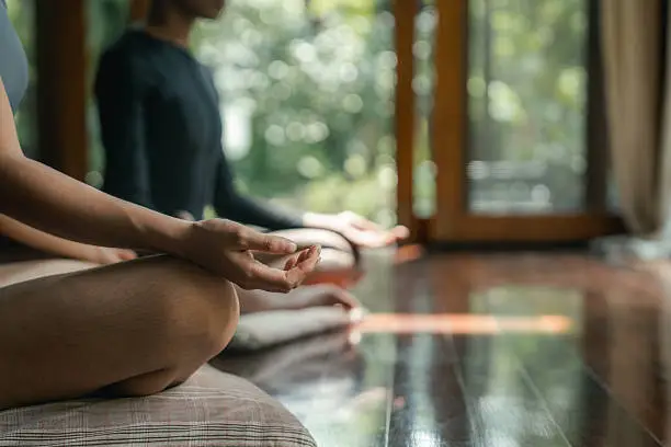 Blog Two women in white activewear sit cross-legged with their eyes closed, practicing mindfulness meditation together in a peaceful, outdoor setting at a wellness resort.