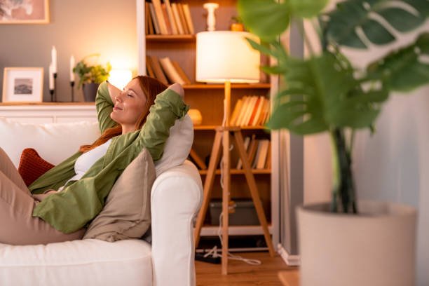 A serene woman with her eyes closed, relaxing on a comfortable sofa with her hands behind her head, enjoying a peaceful, quiet moment that promotes mental health in a sunlit living room.
