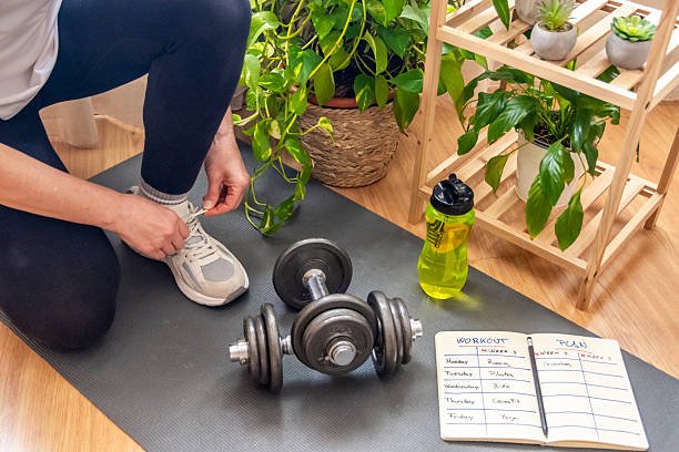 Mature woman tying her shoelaces, preparing for a home workout session with dumbbells, a workout plan, and a refreshing drink, surrounded by houseplants promoting a healthy and active lifestyle Mature woman tying her shoelaces, preparing for a home workout session with dumbbells, a workout plan, and a refreshing drink, surrounded by houseplants promoting a healthy and active lifestyle Weekly Home Workout Routine stock pictures, royalty-free photos & images