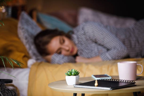 Blog A serene bedroom setting illustrating mindfulness for better sleep, with a journal and succulent in focus on a bedside table and a woman resting peacefully in the background.