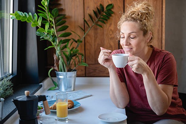 peaceful woman enjoying a hot drink with eyes closed at a breakfast table near a window