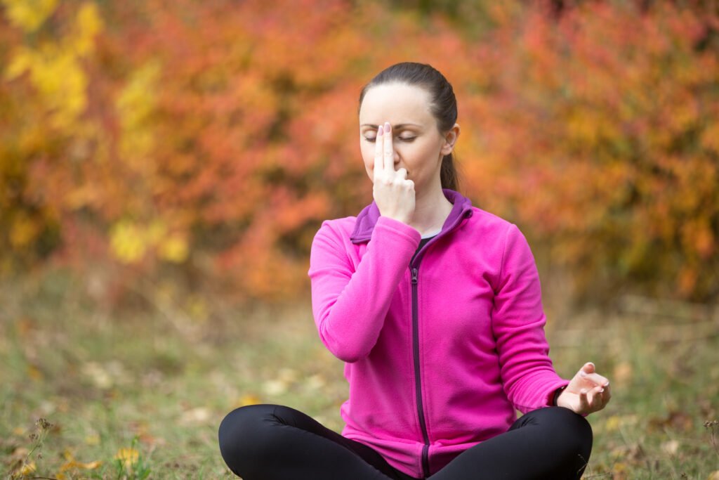 Blog A woman with her eyes closed practices the Nadi Shodhana (alternate nostril breathing) technique for breath awareness in a peaceful autumn park.