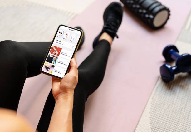 A woman sits on a yoga mat and uses her smartphone to track and adjust your home fitness schedule using a dedicated app.